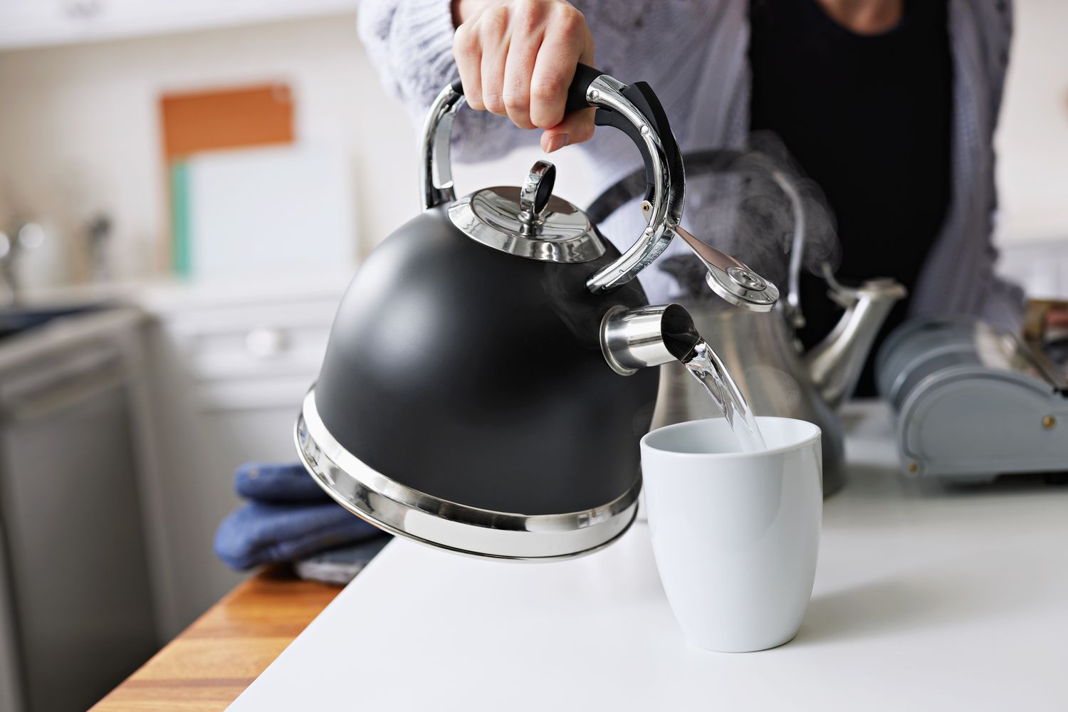 Person pouring hot water from a modern matte-black stovetop kettle into a white mug on a kitchen counter.