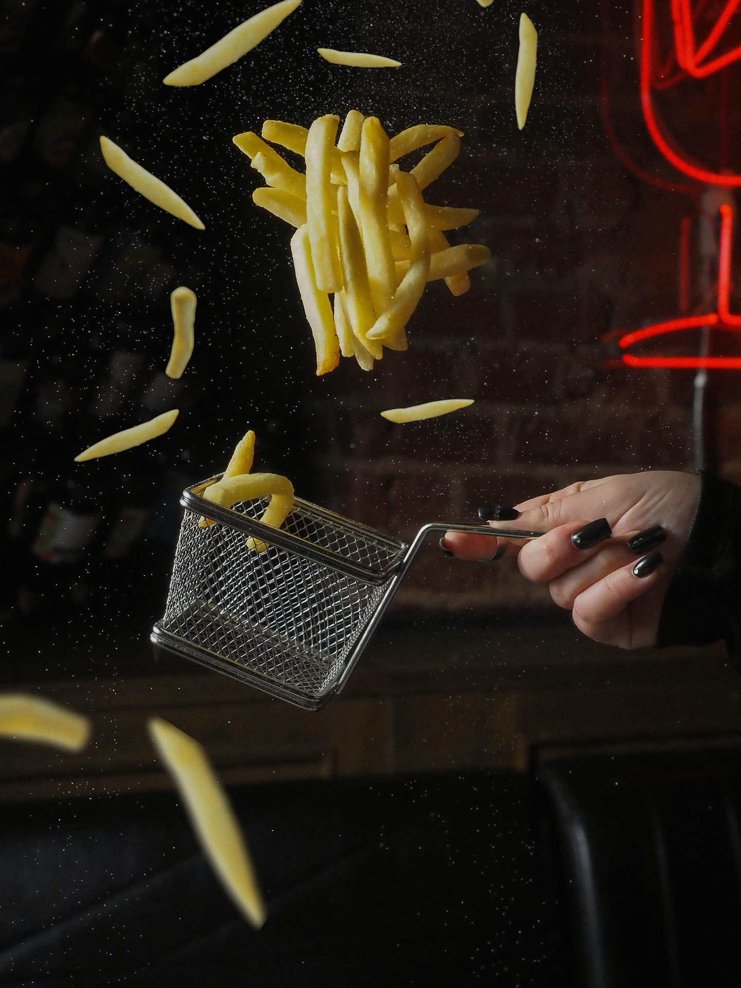 French fries falling into a small fryer basket, captured mid-air before cooking.